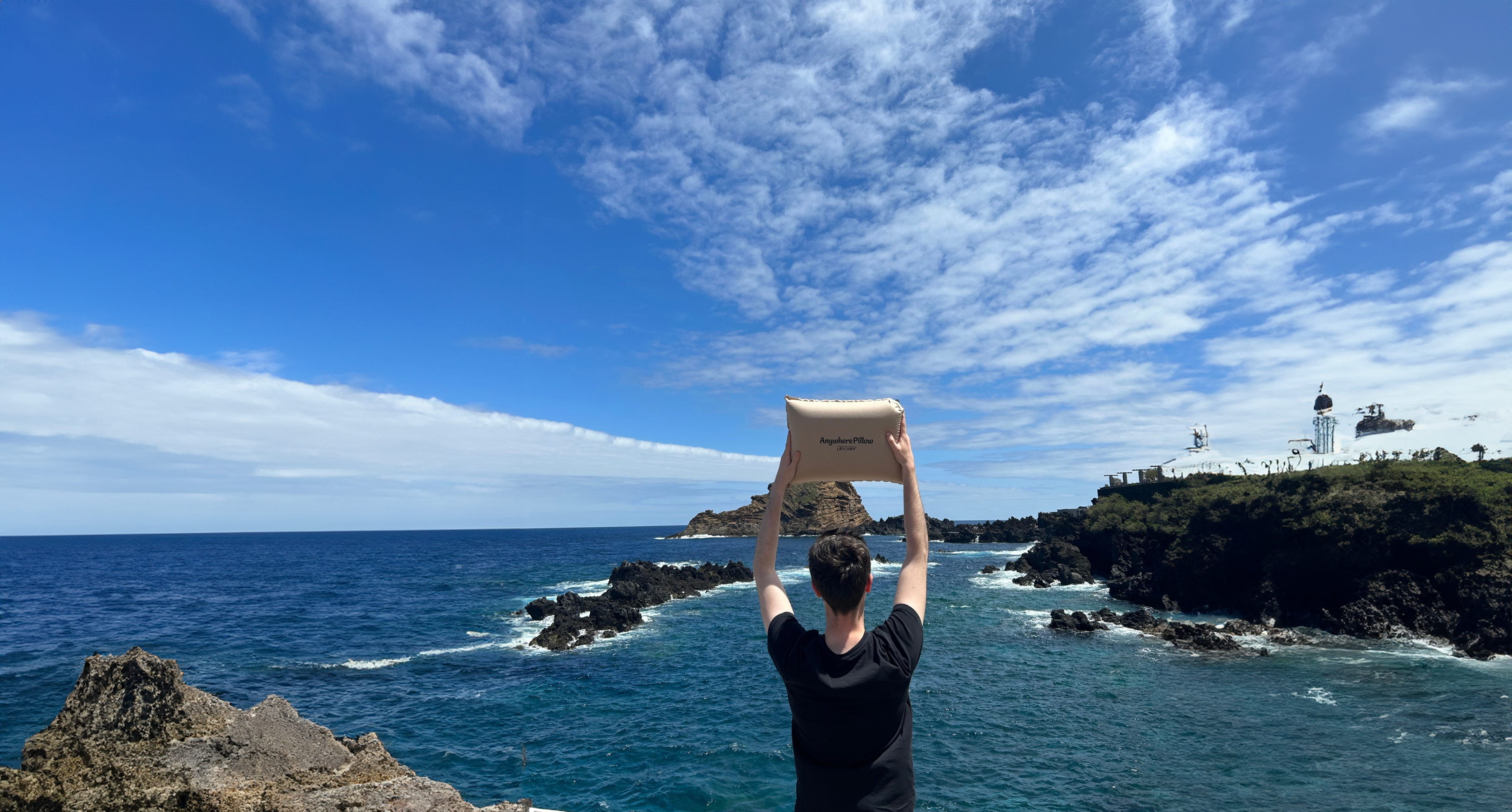 man holding a travel pillow above his head by the sea