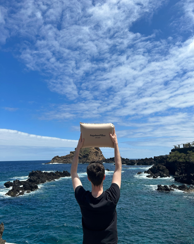 a man holding up a pillow above his head by the sea