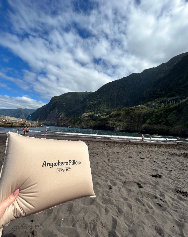 travel pillow held up against the backdrop of a black sand beach and cliffs