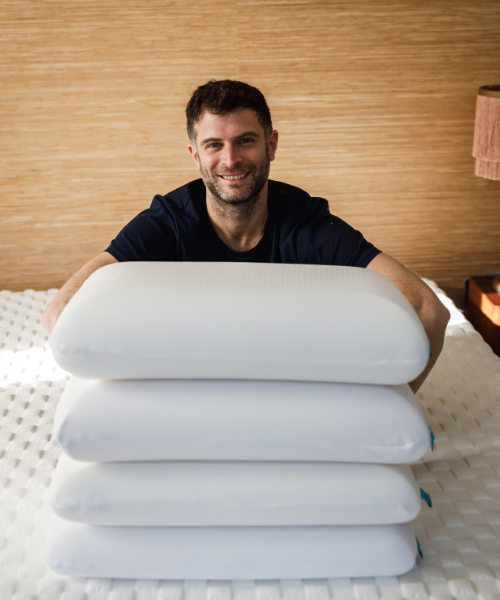 A man sitting behind a pile of four pillows and smiling